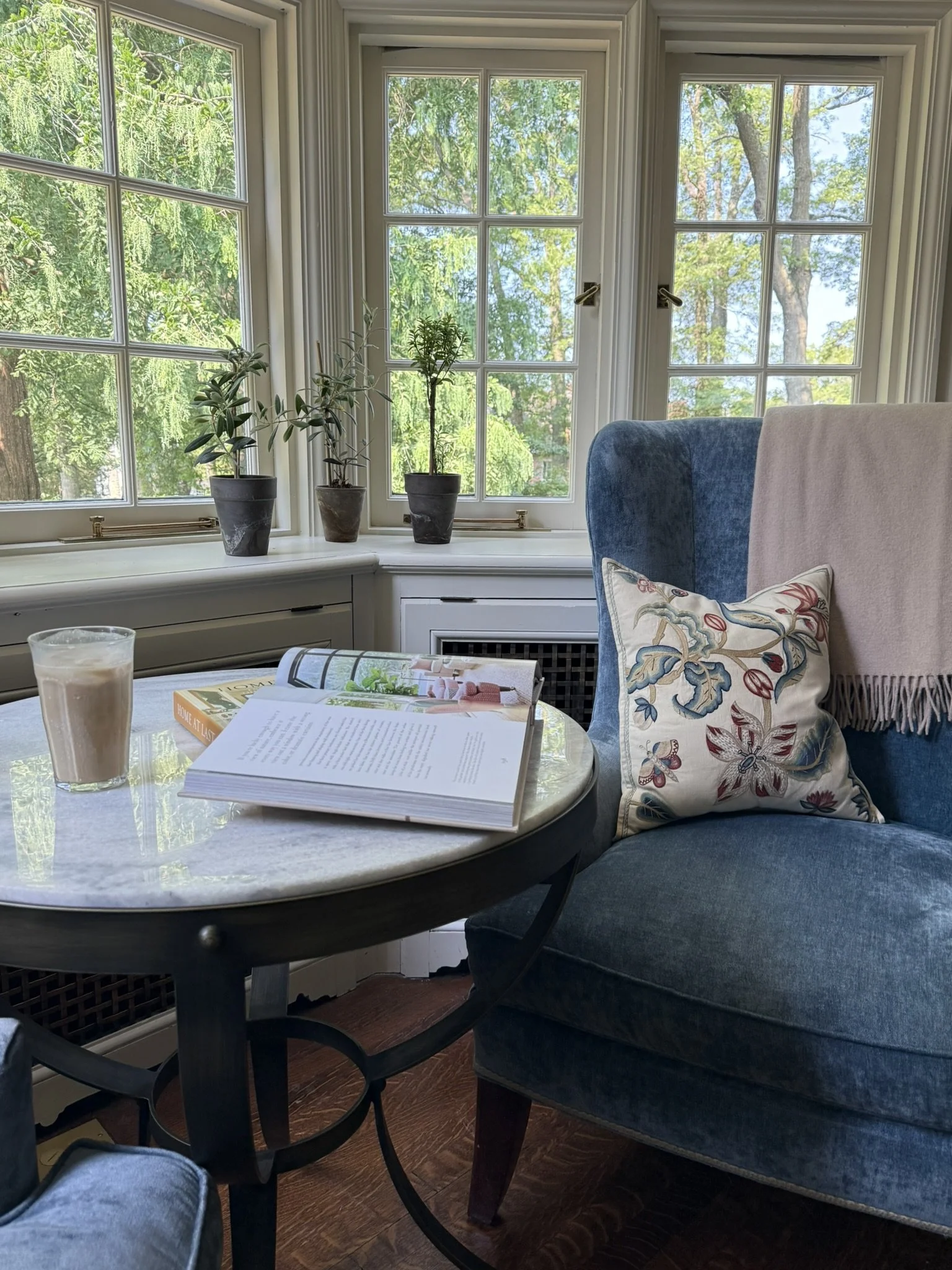 A reading nook with a blue velvet wing chair, an embroidered pillow and a marble side table beside leaded windows.