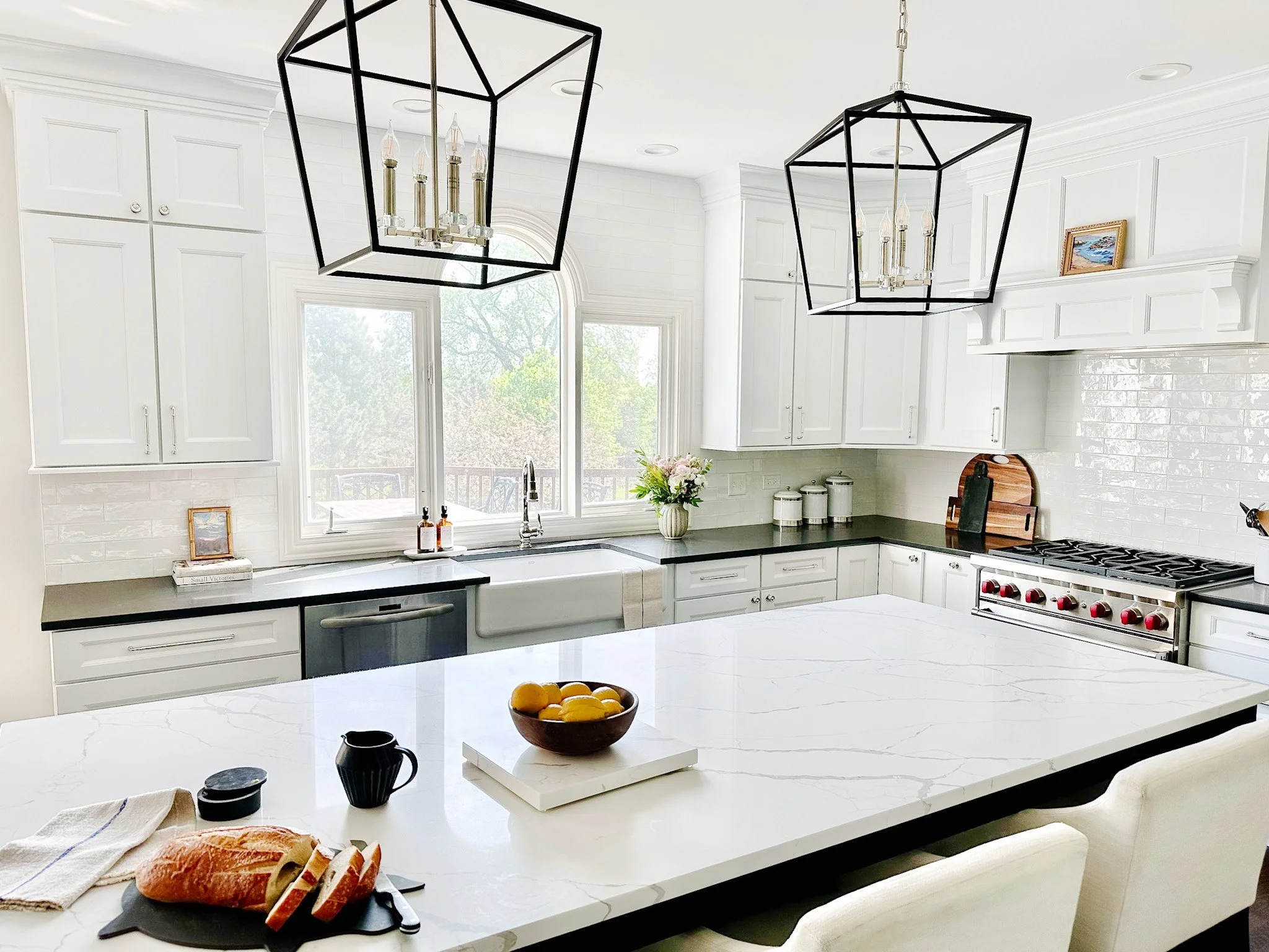 A white kitchen with a large marble island, black countertops and geometric pendant lights.